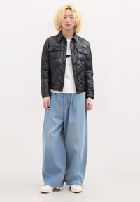 Giacca nera trapuntata con tasche a bottone, abbinata a pantaloni di jeans azzurri larghi e sneakers bianche. L'outfit presenta una vestibilità casual e rilassata.