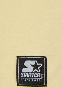 Žltá bavlnená tkanina s čiernym obdĺžnikovým nášivkom, na ktorom je logo "STARTER" a text "BLACK LABEL". Hladká štruktúra, jednoduchý dizajn.