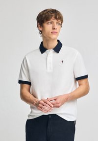 Hvid polo t-shirt med marineblå krave og manchetter, tre-knaps placket og lille rødt logo på venstre bryst. Bæres med mørke bukser.