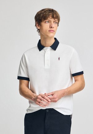 Hvid polo t-shirt med marineblå krave og manchetter, tre-knaps placket og lille rødt logo på venstre bryst. Bæres med mørke bukser.