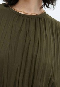 Oliivinvihreä pusero, jossa pystysuoria laskoksia, pyöreä pääntie ja sileä materiaali. Hienovarainen kultainen ketjukoru näkyvissä.