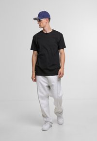 Black cotton T-shirt, gray sweatpants with side seams, white sneakers, and a blue cap. Model stands on a neutral backdrop.