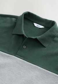 Polo a rayas verdes y grises con cuello, tejido texturizado, un botón negro y etiqueta "NEXT" en el interior del cuello.