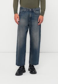Jeans de mezclilla azul de pierna ancha con un acabado desgastado, diseño de cinco bolsillos y cierre de botón. Combinados con zapatos negros de tipo slip-on.
