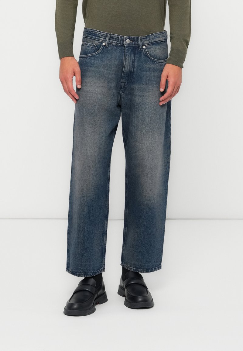 Jeans de mezclilla azul de pierna ancha con un acabado desgastado, diseño de cinco bolsillos y cierre de botón. Combinados con zapatos negros de tipo slip-on.