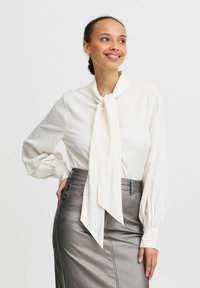 Blouse crème à manches longues avec un col à nouer, dotée de manches bouffantes. Associée à une jupe crayon argent métallique présentant une texture lisse.