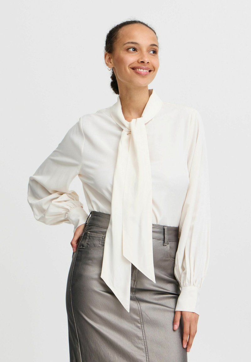 Blouse crème à manches longues avec un col à nouer, dotée de manches bouffantes. Associée à une jupe crayon argent métallique présentant une texture lisse.