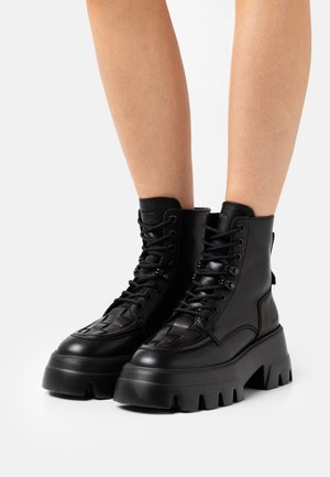 Black chunky platform lace-up boots with thick rugged soles worn on bare legs against a plain white background.