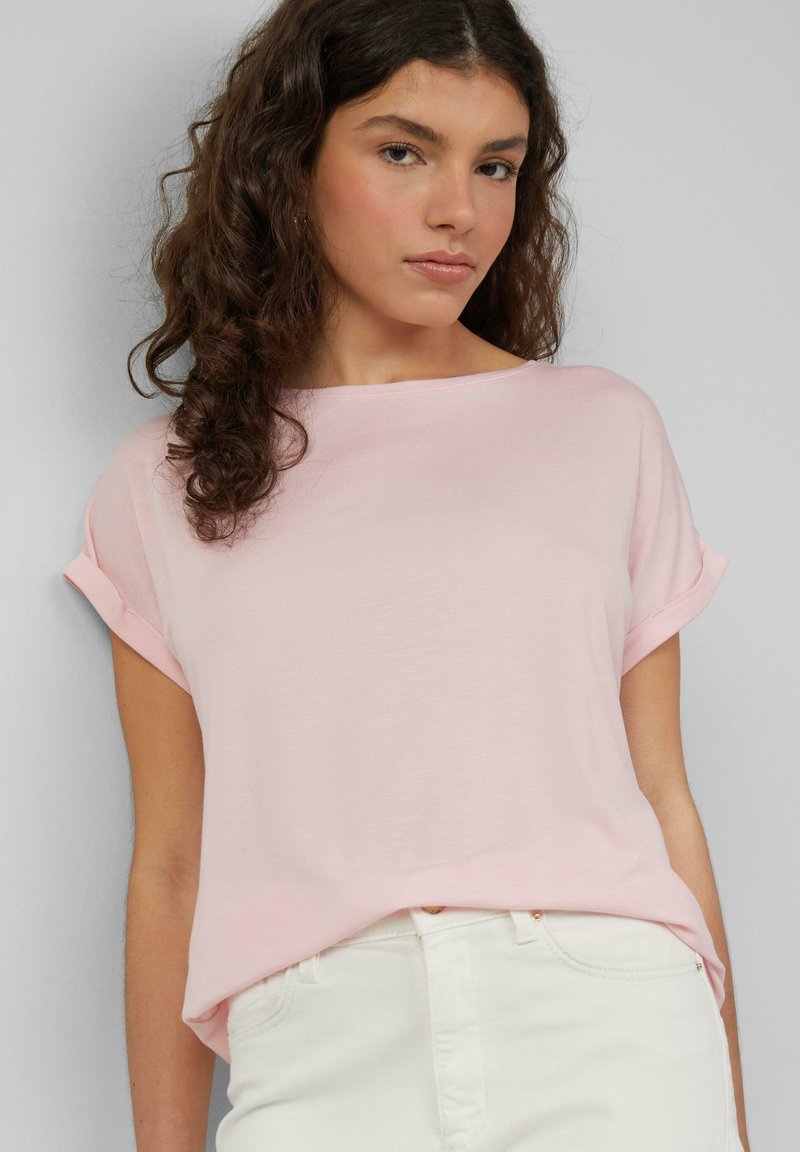 Top corto a maniche corte di colore rosa chiaro con risvolti, realizzato in tessuto morbido. Ha un colletto rotondo e una vestibilità rilassata, abbinato a jeans chiari.