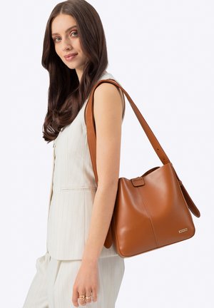 Giovane donna con lunghi capelli castani, indossa un gilet e pantaloni beige a righe sottili, porta una grande borsa a tracolla in pelle marrone.