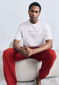 Γκρι βαμβακερό T-shirt με το λογότυπο "TOMMY HILFIGER", συνδυασμένο με κόκκινη φόρμα από μαλακό ύφασμα, με ελαστικά τελειώματα, καθισμένο πάνω σε μια υφή από κρεμ πουφ.