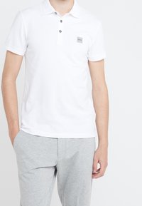 Valkoinen polo-paita, jossa on pehmeä pinta, kolmesta napista koostuva nappilista ja pieni harmaa logopatchi rinnassa. Yhdistetty vaaleanharmaisiin housuihin.