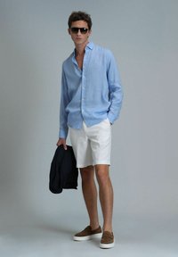Hellblaues Leinenhemd, weiße Shorts, braune Loafers, hält eine dunkelblaue Jacke, trägt Sonnenbrille; lockere Passform und lässige Kleidung.