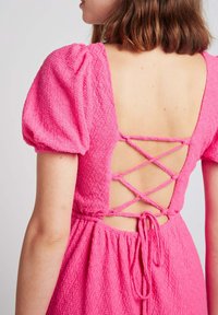 Robe rose texturée avec des manches bouffantes et un dos lacé. Le matériau présente un motif subtil ; le dos est décolleté et se noue pour l'ajustement.