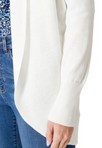 Cardigan blanc léger en maille côtelée avec un devant drapé, des manches ajustées et une texture douce, associé à un jean en denim bleu.