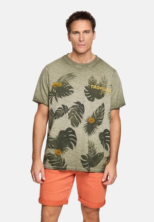 Hombre con camiseta de color verde oliva con estampado tropical de flores amarillas y pantalones cortos coral enrollados, de pie contra un fondo claro.