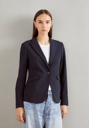 Marineblazer in maßgeschneidertem Design, mit zwei vorderen Taschen, einem einzigen Knopfverschluss und glattem Stofffinish, getragen über einem weißen Oberteil.