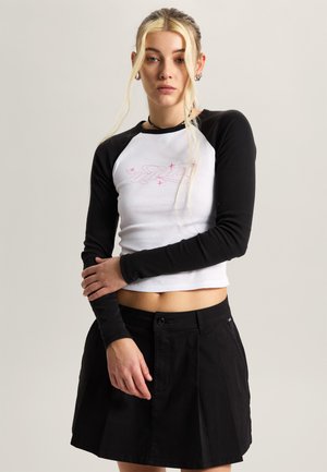 Młoda kobieta z długimi blond włosami nosi biały crop top z czarnymi rękawami i czarną plisowaną spódnicę, stoi na tle jednolitego tła.