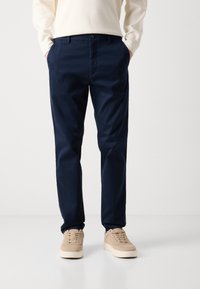 Navy blue chinos made of cotton. Tailored fit with a smooth texture, zip fly, and front pockets. Paired with beige sneakers.
