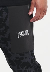 Černé tepláky s texturovaným vzorem, disponující zipovou kapsou a vyšitým nápisem "PEGADOR" na boku.