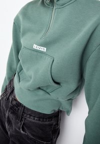 Felpa verde con colletto con zip, tasca frontale e patch con il logo Levi's. Abbinata a jeans neri, che mostrano un fit rilassato.