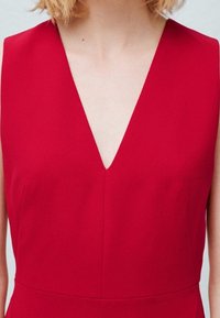 Robe rouge avec un décolleté en V, sans manches, texture de tissu lisse et silhouette ajustée. Présente un détail de couture subtil au niveau de la poitrine.