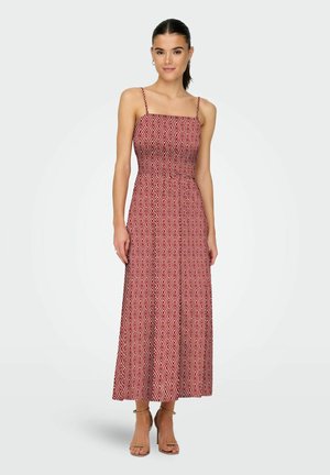 Frau trägt ein ärmelloses, knöchellanges rotes Kleid mit weißen geometrischen Rautenmuster und dünnen Schulterträgern, kombiniert mit beigen High Heels.