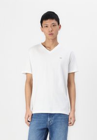 T-shirt blanc à col en V fabriqué en coton doux, avec des manches courtes et un petit logo sur la poitrine. Porté avec un jean bleu.