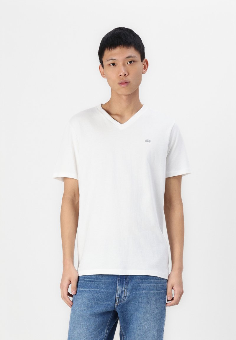 T-shirt blanc à col en V fabriqué en coton doux, avec des manches courtes et un petit logo sur la poitrine. Porté avec un jean bleu.