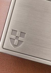 Produkt z matowego metalu w kształcie prostokąta, z wytłaczanym logo z elementami geometrycznymi. Tło stanowi teksturowana brązowa powierzchnia.