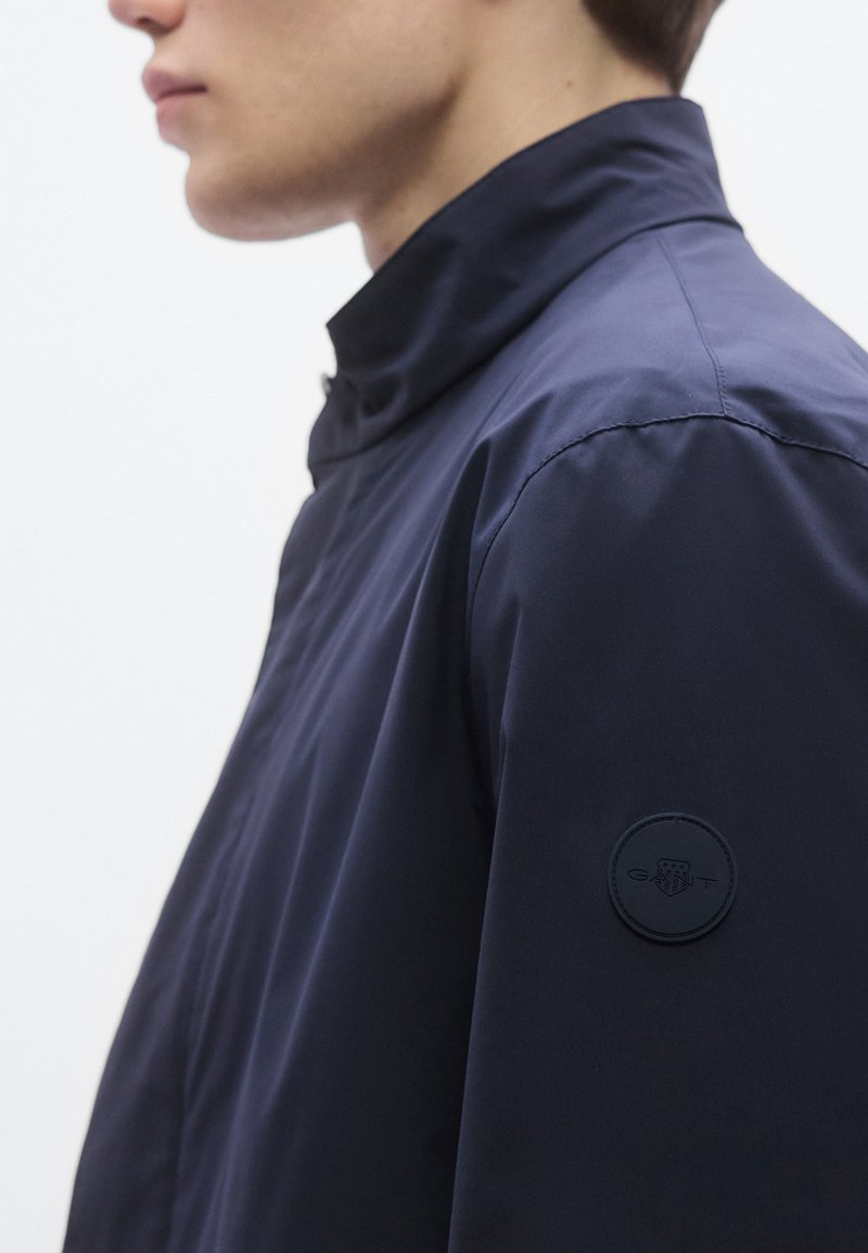 Navy blue jacket with a high collar, featuring smooth texture and a circular black logo patch on the upper arm.