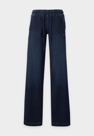 Femme debout les mains dans le dos, portant un top court noir Diesel, une veste bleu marine et un pantalon large bleu marine sur un fond uni.