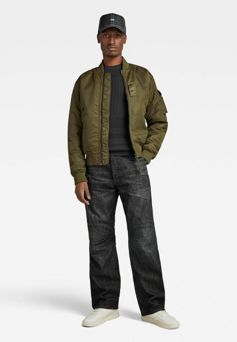 Un homme portant une casquette noire, une veste bomber olive, un pull noir à côtes, un jean foncé et des baskets blanches, se tenant avec une main dans la poche.