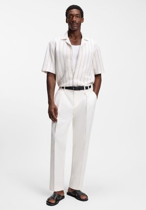 Mann steht vor einfarbigem Hintergrund, trägt ein weiß gestreiftes Kurzarmhemd über einem weißen Tanktop, weiße maßgeschneiderte Hose, schwarzen Gürtel und schwarze Sandalen.