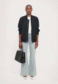 Veste bomber noire, t-shirt blanc, jeans évasés bleu clair, talons blancs avec des accents noirs, tenant un sac à main en textile noir.