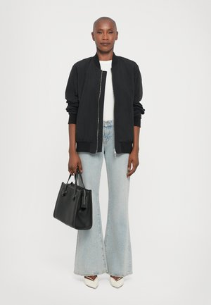 Giacca bomber nera, t-shirt bianca, jeans a campana blu chiaro, tacchi bianchi con accenti neri, in mano una borsa nera con texture.