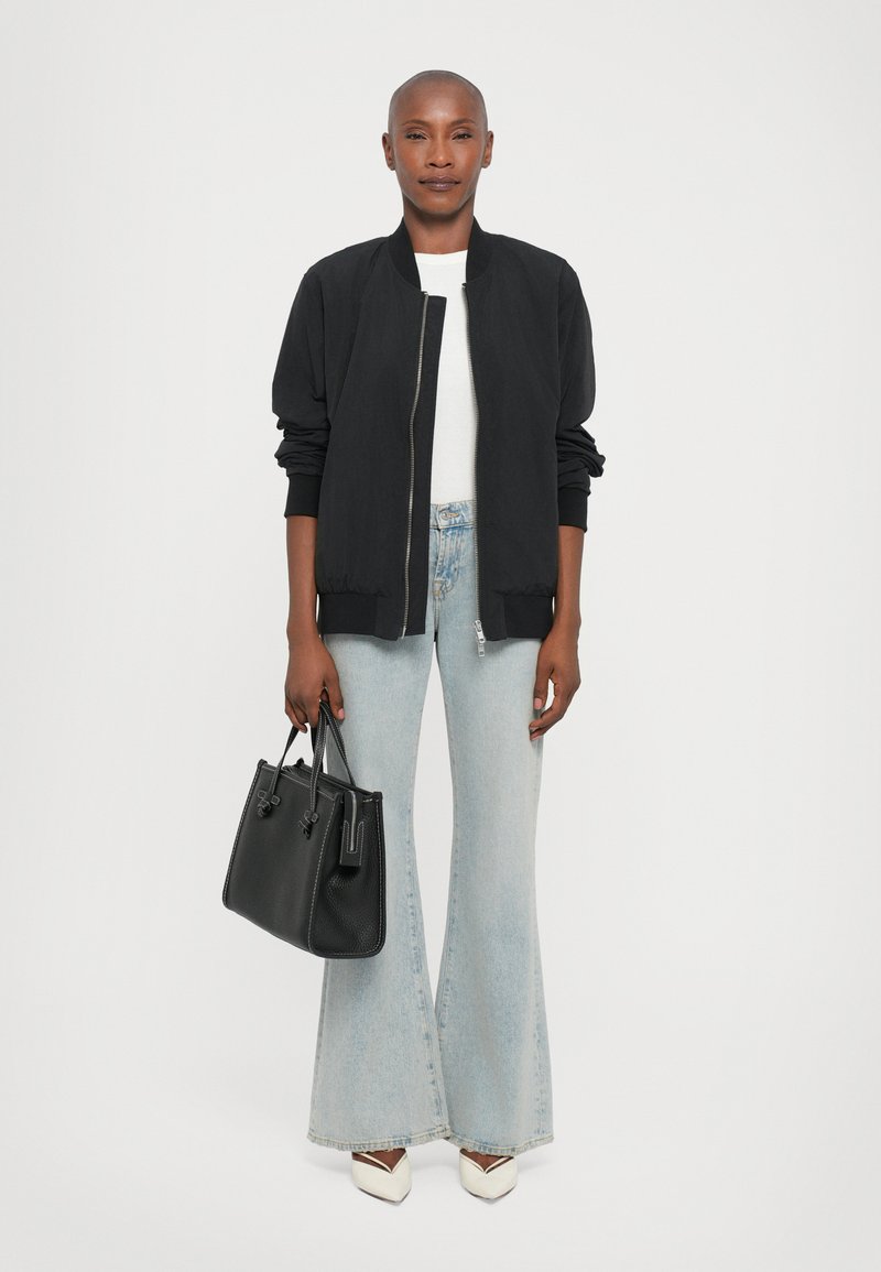 Veste bomber noire, t-shirt blanc, jeans évasés bleu clair, talons blancs avec des accents noirs, tenant un sac à main en textile noir.