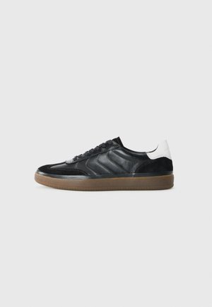 Black low-top sneaker with brown sole, lace-up front, chevron side details, and white heel patch, shown in right profile on gray background.