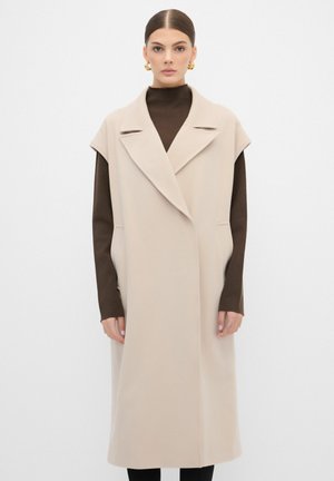 Beige ärmelloser Mantel mit weitem Kragen, aus strukturiertem Stoff gefertigt. Besitzt seitliche Taschen und eine lockere Passform, über einem braunen Oberteil getragen.