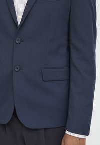 Tengerészkék zakó, textúrált anyagból készült, két gombbal, mellzsebbel és alatta kontrasztos csíkozott ingmandzsettával.