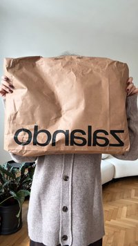 Henkilö beige neulepuserossa pitää suurta ruskeaa paperista Zalando-ostoskassia kasvonsa edessä olohuoneessa, jossa on kasvi ja valkoinen sohva.