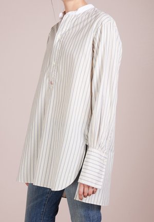 Kvinde iført løs, langærmet, hvid skjorte med tynde blå lodrette striber og brede manchetter, kombineret med blå jeans, mod en neutral baggrund.