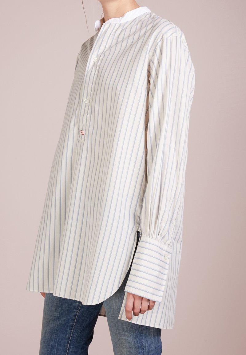 Femme portant une chemise blanche ample à manches longues avec de fines rayures verticales bleues et de larges poignets, associée à un jean bleu, sur un fond neutre.