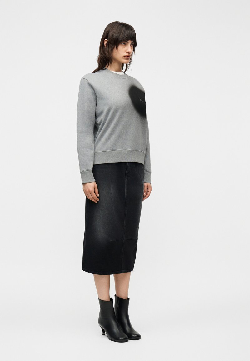 Femme debout portant un sweat-shirt gris avec une tache noire, une jupe midi noire et des bottines à talons noires, sur un fond blanc uni.