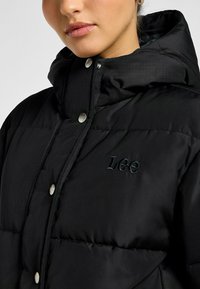 Sort pufferjakke med quiltet design, høj krave og blanke sølvknapper. Har et broderet "Lee"-logo på brystet.