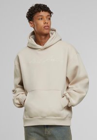 Beige hættetrøje med frontlomme, med broderet "Karl Kani" logo i hvid. Blød tekstur, løs pasform og ribbede manchetter.