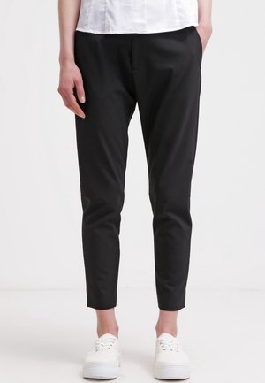Personne portant un pantalon noir slim longueur cheville, des baskets blanches et une chemise blanche, debout devant un fond uni.