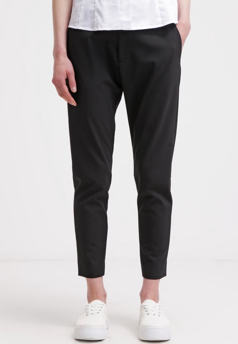 Personne portant un pantalon noir slim longueur cheville, des baskets blanches et une chemise blanche, debout devant un fond uni.