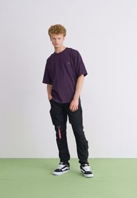 Čierne cargo nohavice s bočnými vreckami, fialové oversized tričko a čierno-biele tenisky. Model stojí na svetlozelenej podlahe.