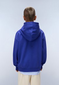 Sudadera azul real con una gran capucha, hombros caídos y puños acanalados. Tejido suave, corte relajado, superpuesta a una camisa blanca.